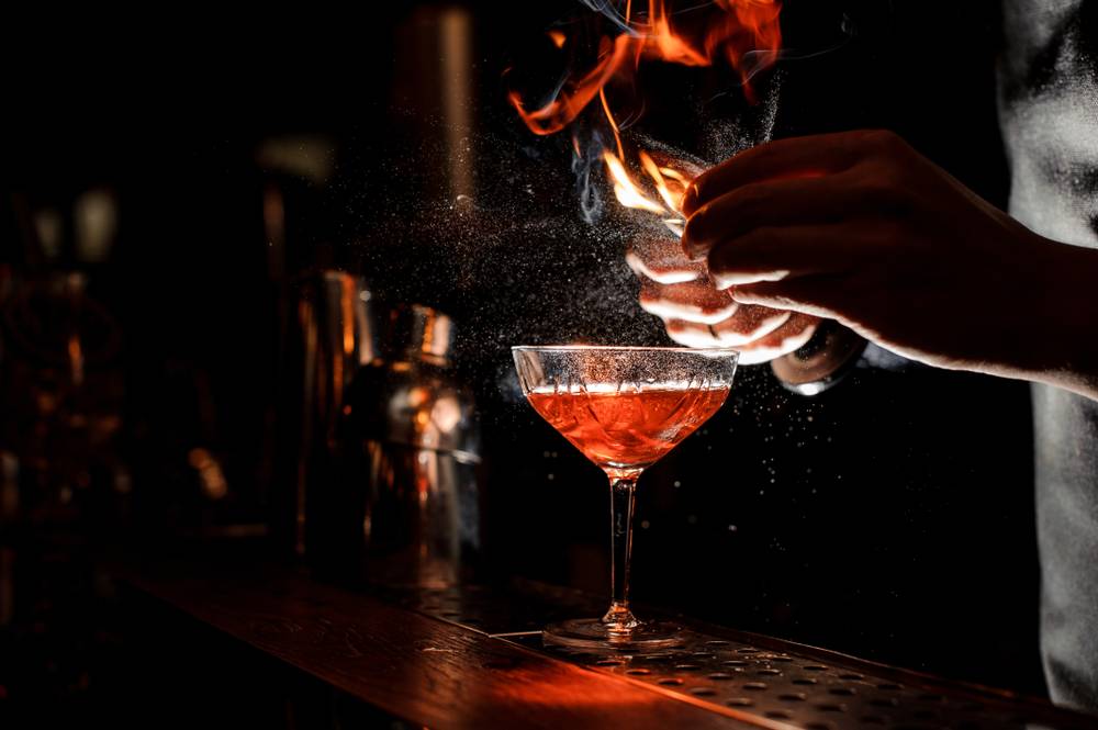 Tsim Sha Tsui Bar Bartender`s hands sprinkling the juice into the cocktail glass filled with alcoholic drink and making a smoky note on the dark background